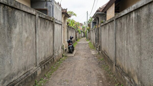 A very narrow dirt alleyway lined with tall concrete walls in a residential area, illustrating road access mistakes buying property in Bali.